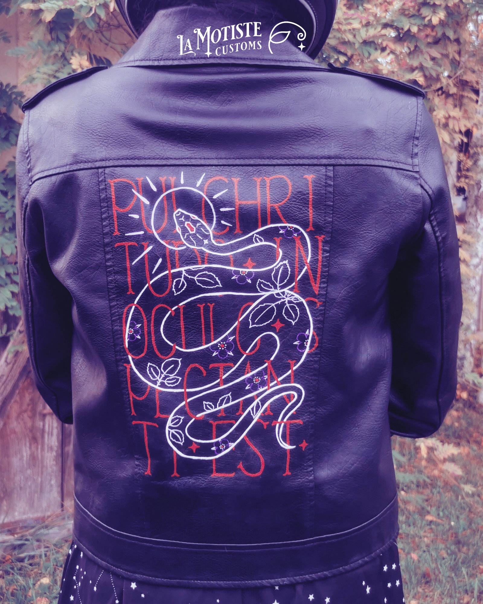Gros plan sur le dos d'une veste en simili cuir peinte par la motiste. Le motif représente les contours d'un serpent en blanc, une auréole violet clair autour de sa tête, et des feuilles et fleurs de ronces violet et rouge en motifs sur son corps. Le serpent est entremêlé dans la phrase Pulchritudo in oculo spectanti est, écrite en rouge en caractères fin majuscule.