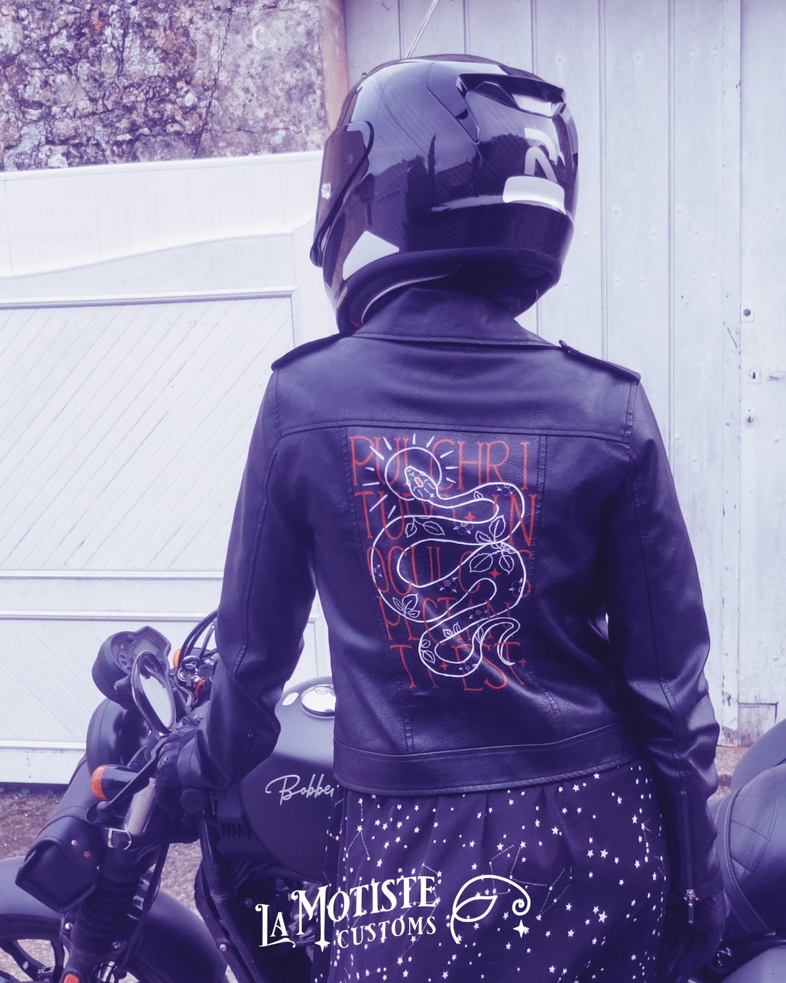 Photo d'une personne casquée de dos, portant une veste en simili cuir peinte par la motiste. Le motif représente les contours d'un serpent en blanc, une auréole violet clair autour de sa tête, et des feuilles et fleurs de ronces violet et rouge en motifs sur son corps. Le serpent est entremêlé dans la phrase Pulchritudo in oculo spectanti est, écrite en rouge en caractères fin majuscule.