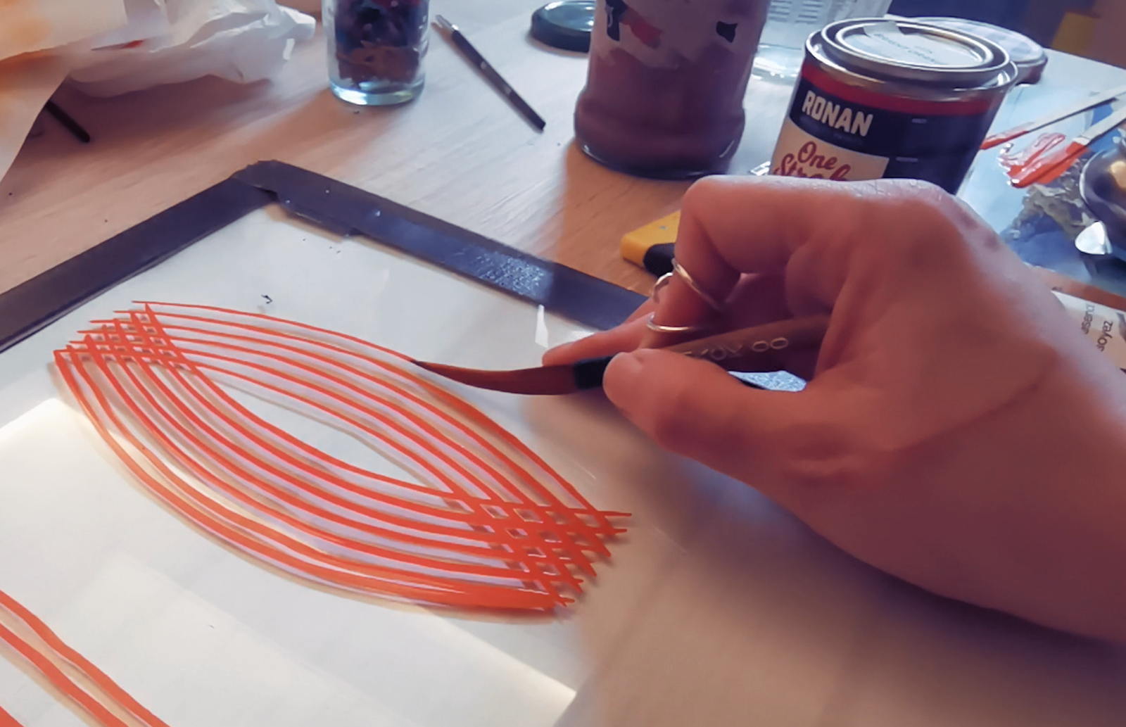 Photo d'une main tenant un pinceau de pinstriping et peignant un motif composé de courbes en C parallèles les unes aux autres, croisant un deuxième groupe de courbes en C du sens opposé