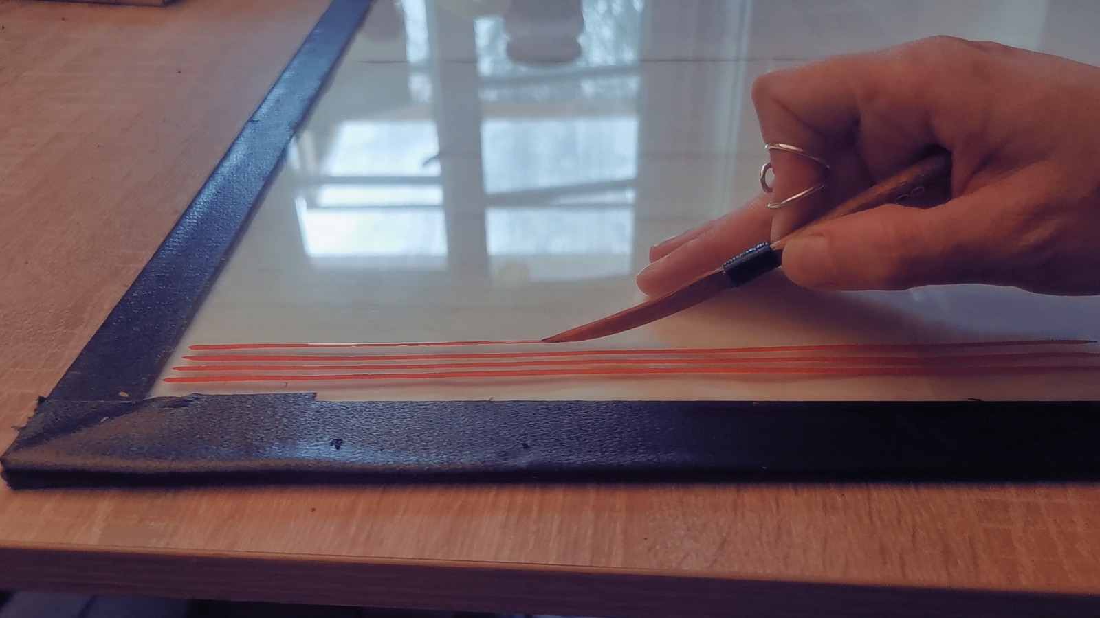 Photo en gros plan d'une main tenant un pinceau de pinstriping et peignant des lignes droites oranges sur une plaque en verre