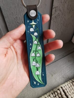 Photo du porte clés Muguet. Le cuir est bleu-vert foncé, le motif est un brin de muguet peint dans un style légèrement bd, sur fond étoilé
