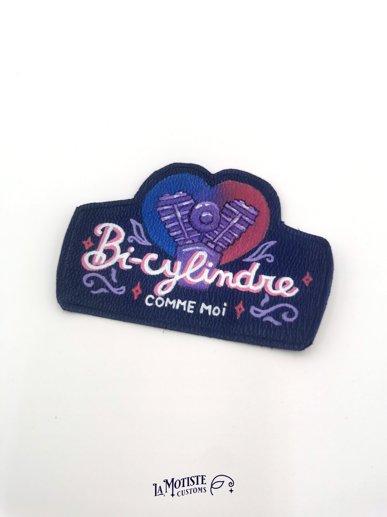 Patch en cuir peint à la main. Un moteur bicylindre est peint sur un fond en forme de coeur au dégradé couleurs du drapeau bisexuel : bleu violet et rose. Devant l'illustration le texte "bi-cylindre" écrit dans une typographie un peu rétro