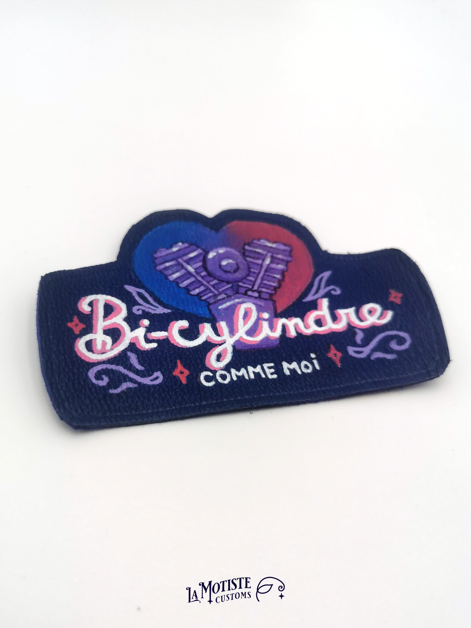Patch en cuir peint à la main. Un moteur bicylindre est peint sur un fond en forme de coeur au dégradé couleurs du drapeau bisexuel : bleu violet et rose. Devant l'illustration le texte "bi-cylindre" écrit dans une typographie un peu rétro
