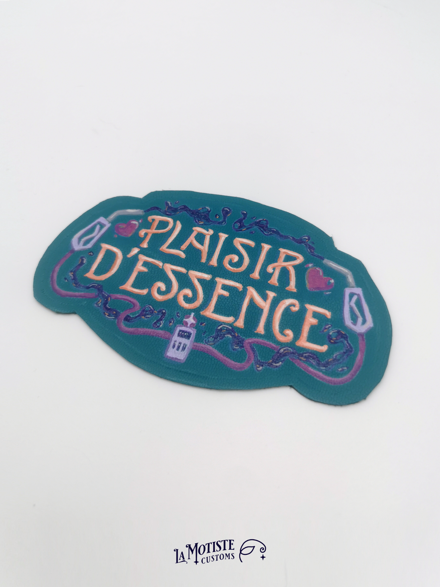 Patch en cuir peint à la main Plaisir d'Essence. Le cuir est bleu-vert, le texte dans une typographie art nouveau orang, entoutée de deux pompes à essence violettes et d'essence violette foncé
