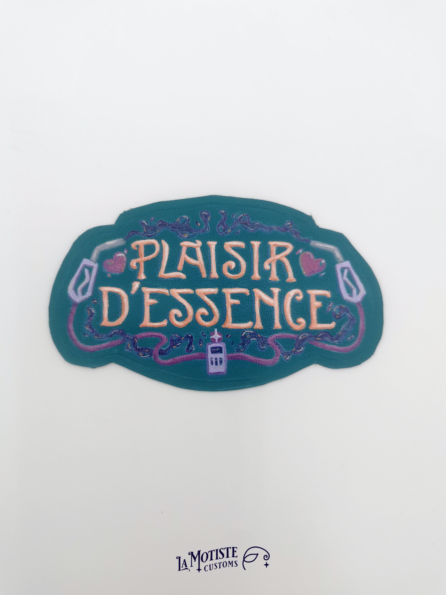 Patch en cuir peint à la main Plaisir d'Essence. Le cuir est bleu-vert, le texte dans une typographie art nouveau orang, entoutée de deux pompes à essence violettes et d'essence violette foncé