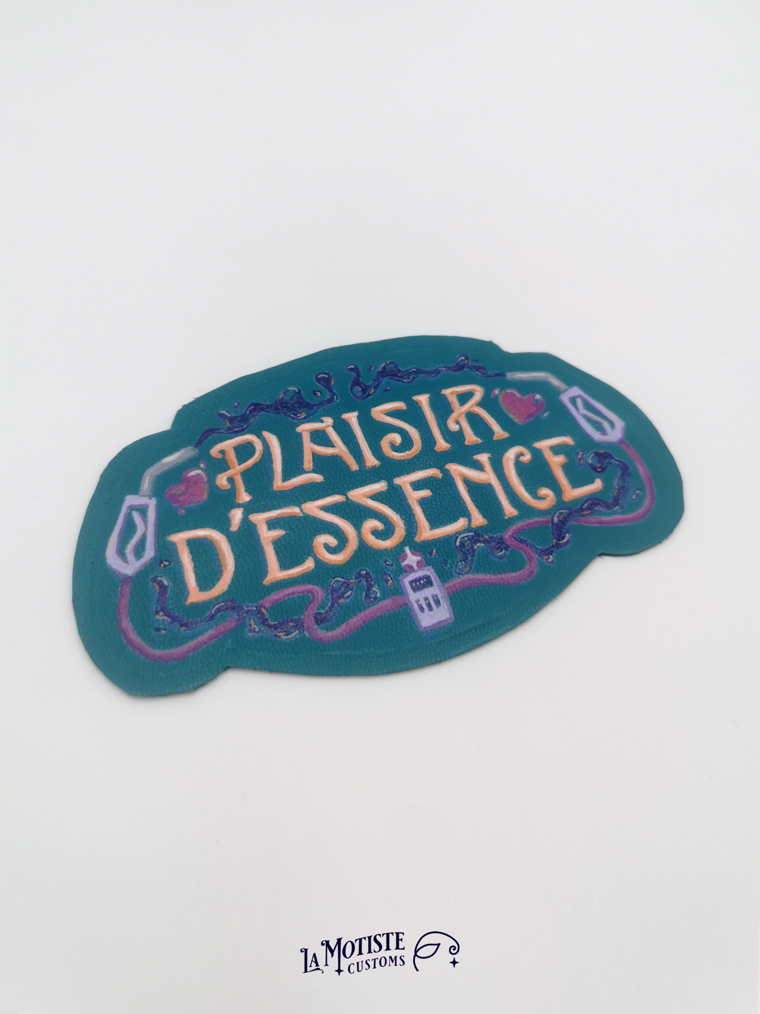 Patch en cuir peint à la main Plaisir d'Essence. Le cuir est bleu-vert, le texte dans une typographie art nouveau orang, entoutée de deux pompes à essence violettes et d'essence violette foncé