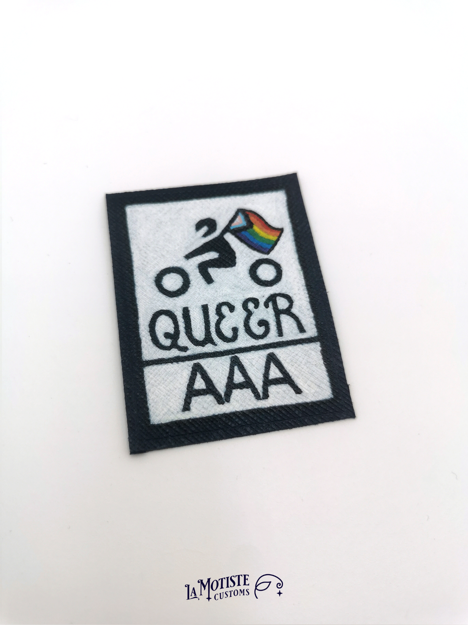 Patch en cuir Queer AAA qui s'inspire du design du logo de certification de résistance à l'abrasion des équipements moto. Le petit motard tiens un drapeau lgbtqia+ inclusif