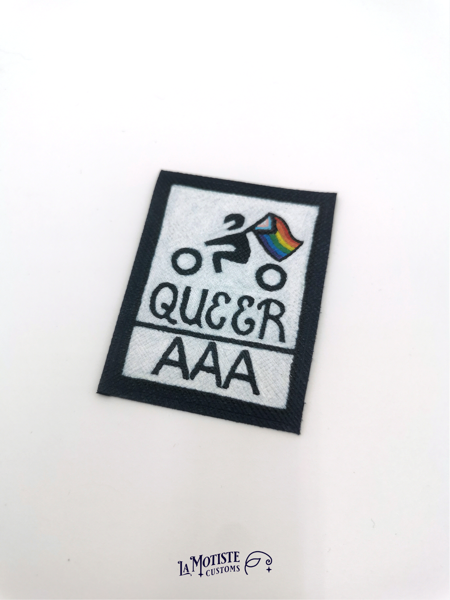Patch en cuir Queer AAA qui s'inspire du design du logo de certification de résistance à l'abrasion des équipements moto. Le petit motard tiens un drapeau lgbtqia+ inclusif