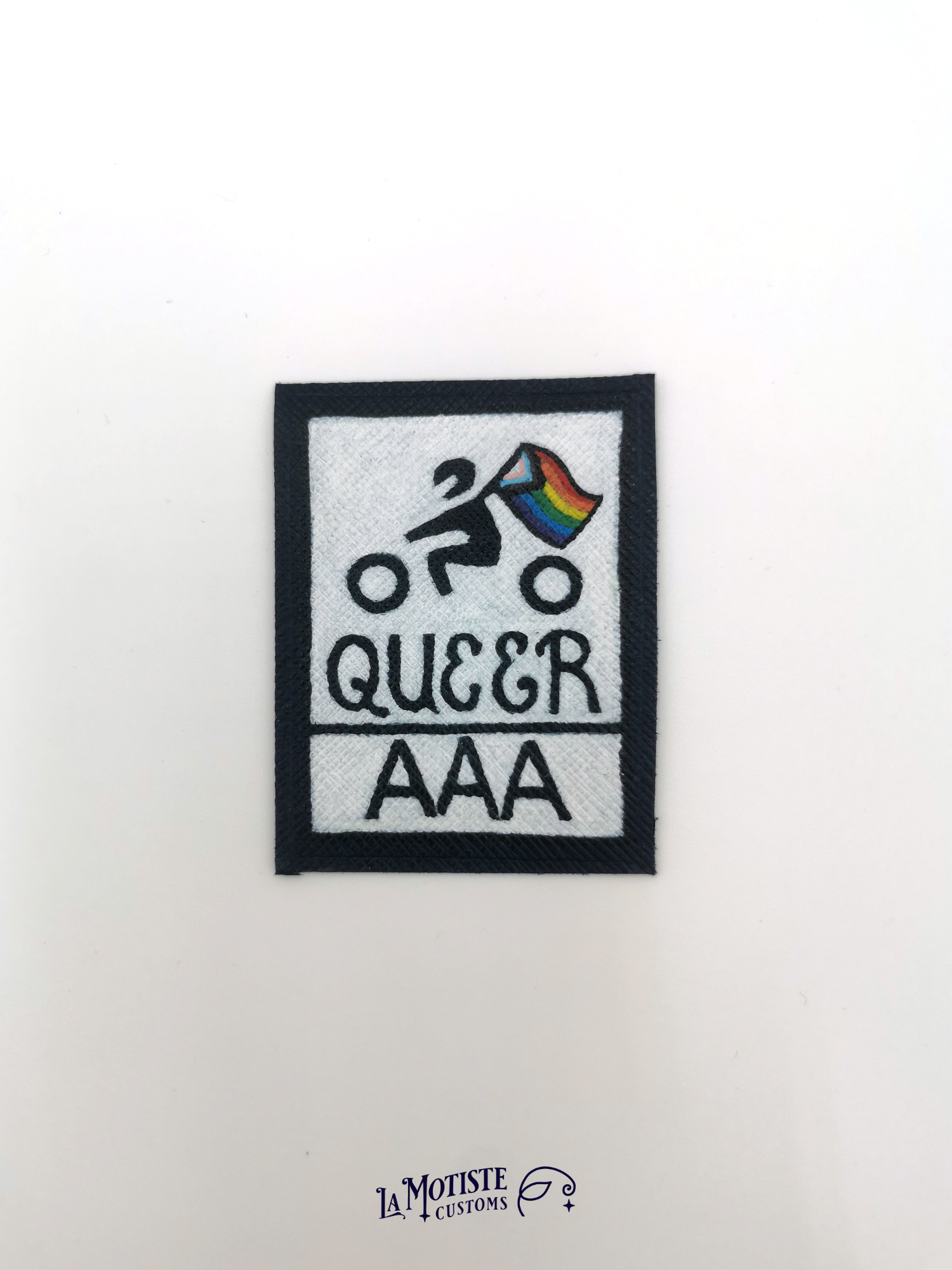 Patch en cuir Queer AAA qui s'inspire du design du logo de certification de résistance à l'abrasion des équipements moto. Le petit motard tiens un drapeau lgbtqia+ inclusif