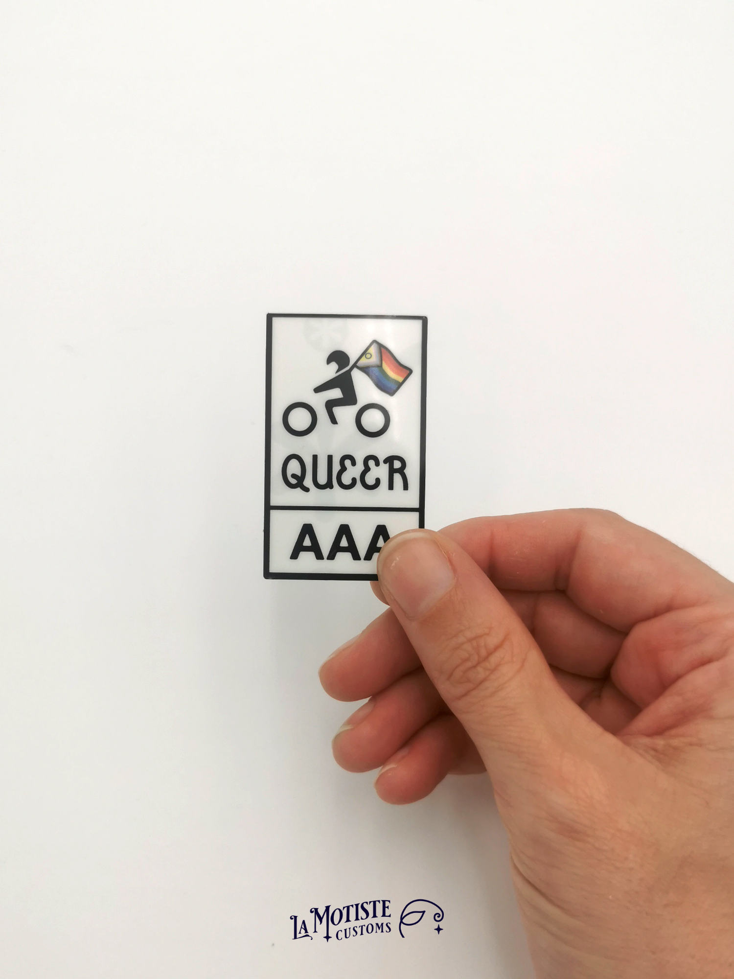 Stickers pelliculé en plastique recyclé Queer AAA. l'icône de motard porte un drapeau lgbtqia+ inclusif, texte Queer AAA, porté à la main pour avoir une idée de la taille