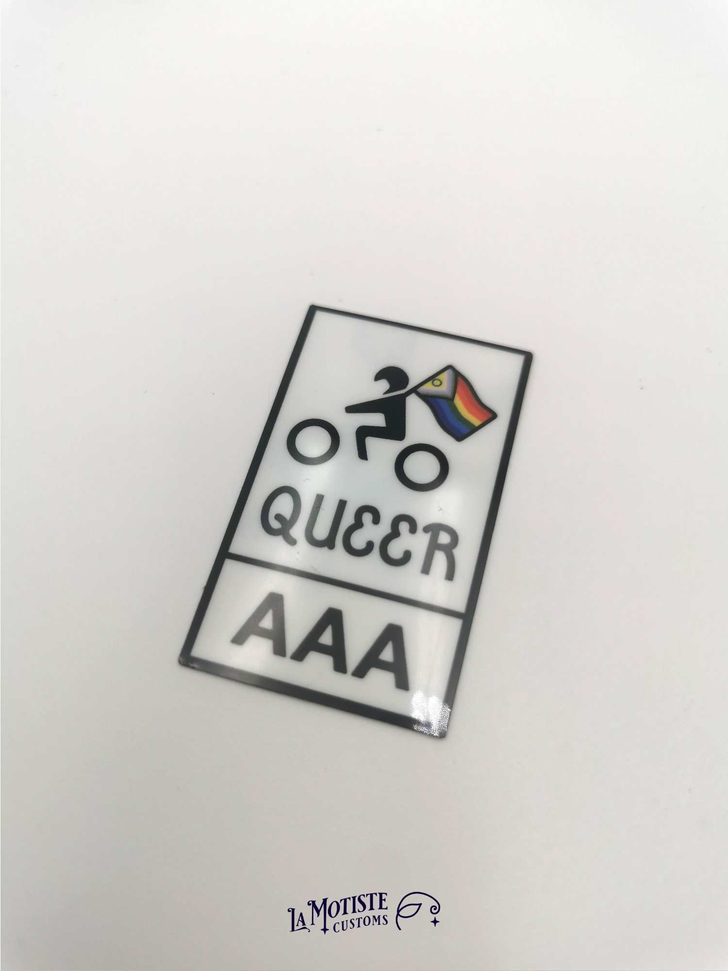 Stickers pelliculé en plastique recyclé Queer AAA. l'icône de motard porte un drapeau lgbtqia+ inclusif, texte Queer AAA, porté à la main pour avoir une idée de la taille