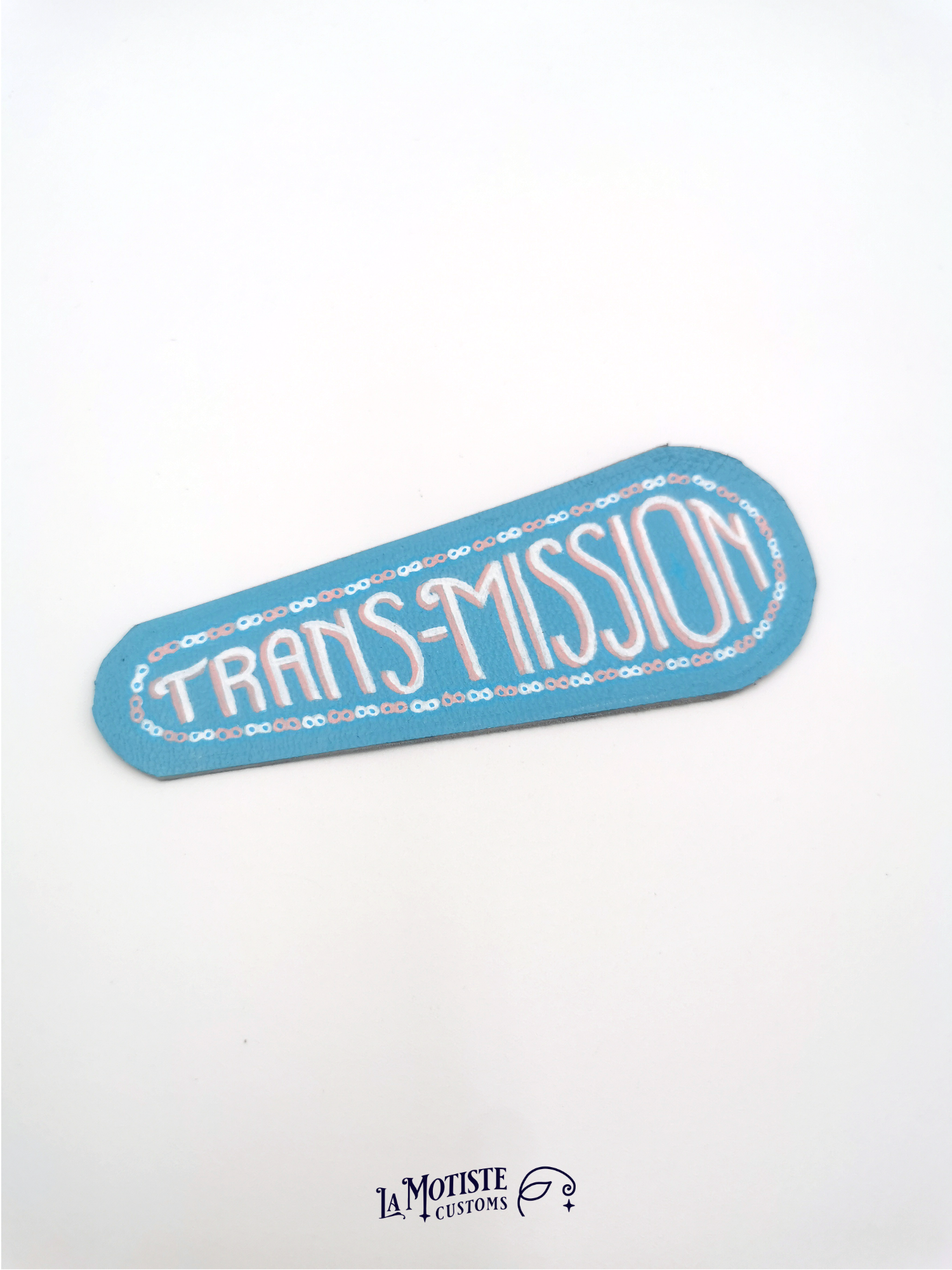 Patch en cuir peint à la main trans-misison. Le cuir est bleu clair, un motif de chaîne de transmission au maillons alternés rose et blanc, et à l'intérieur le mot trans-mission peint dans une écriture art déco