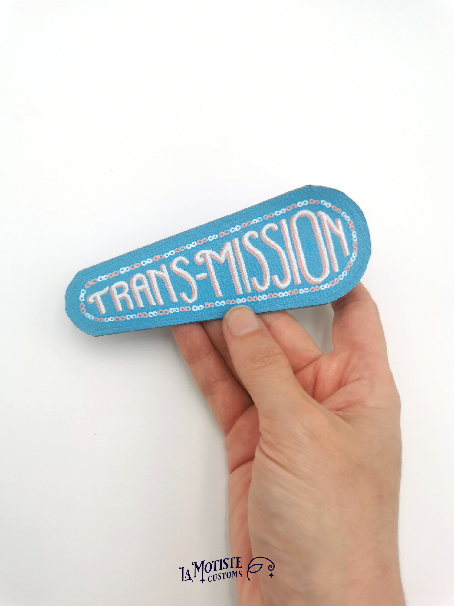 Patch en cuir peint à la main trans-misison. Le cuir est bleu clair, un motif de chaîne de transmission au maillons alternés rose et blanc, et à l'intérieur le mot trans-mission peint dans une écriture art déco